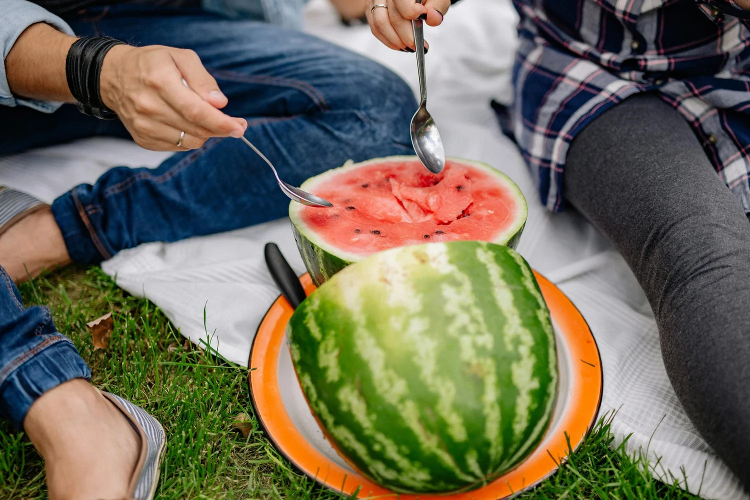 Two people scooping watermelon with spoons on a picnic blanket