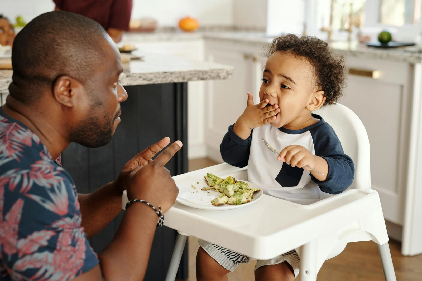A father sitting with his laughing toddler in a high chair at mealtime
