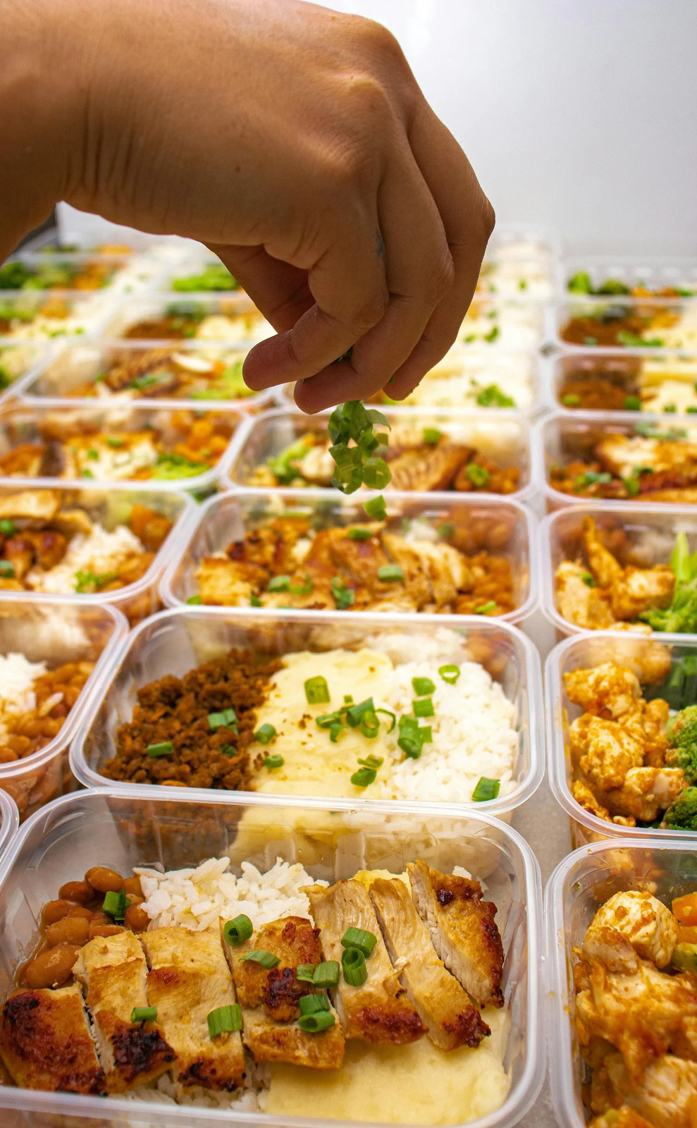 Scallions being sprinkled over rows of prepared meals in containers