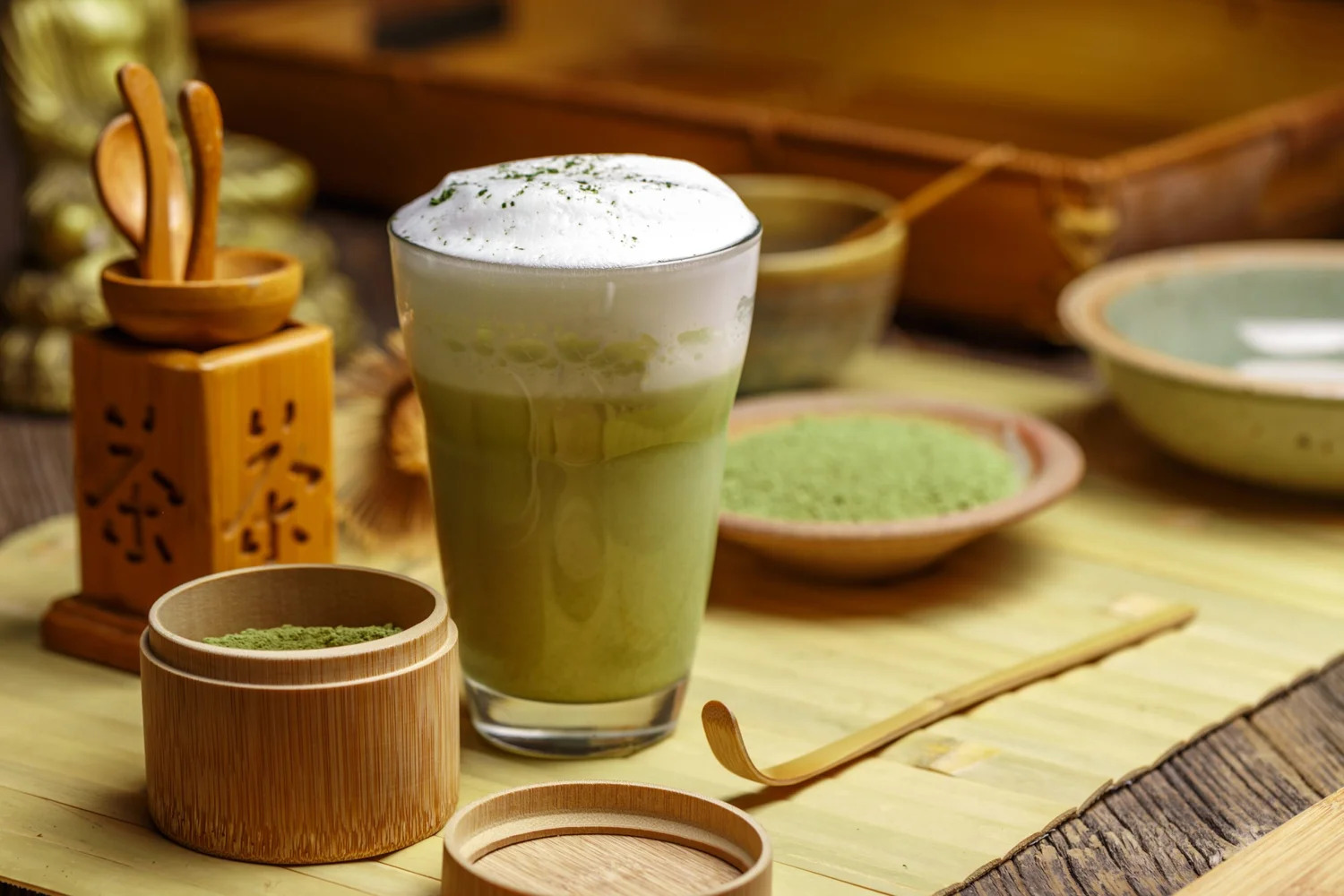 A matcha latte beside bowls of matcha powder and a bamboo whisk