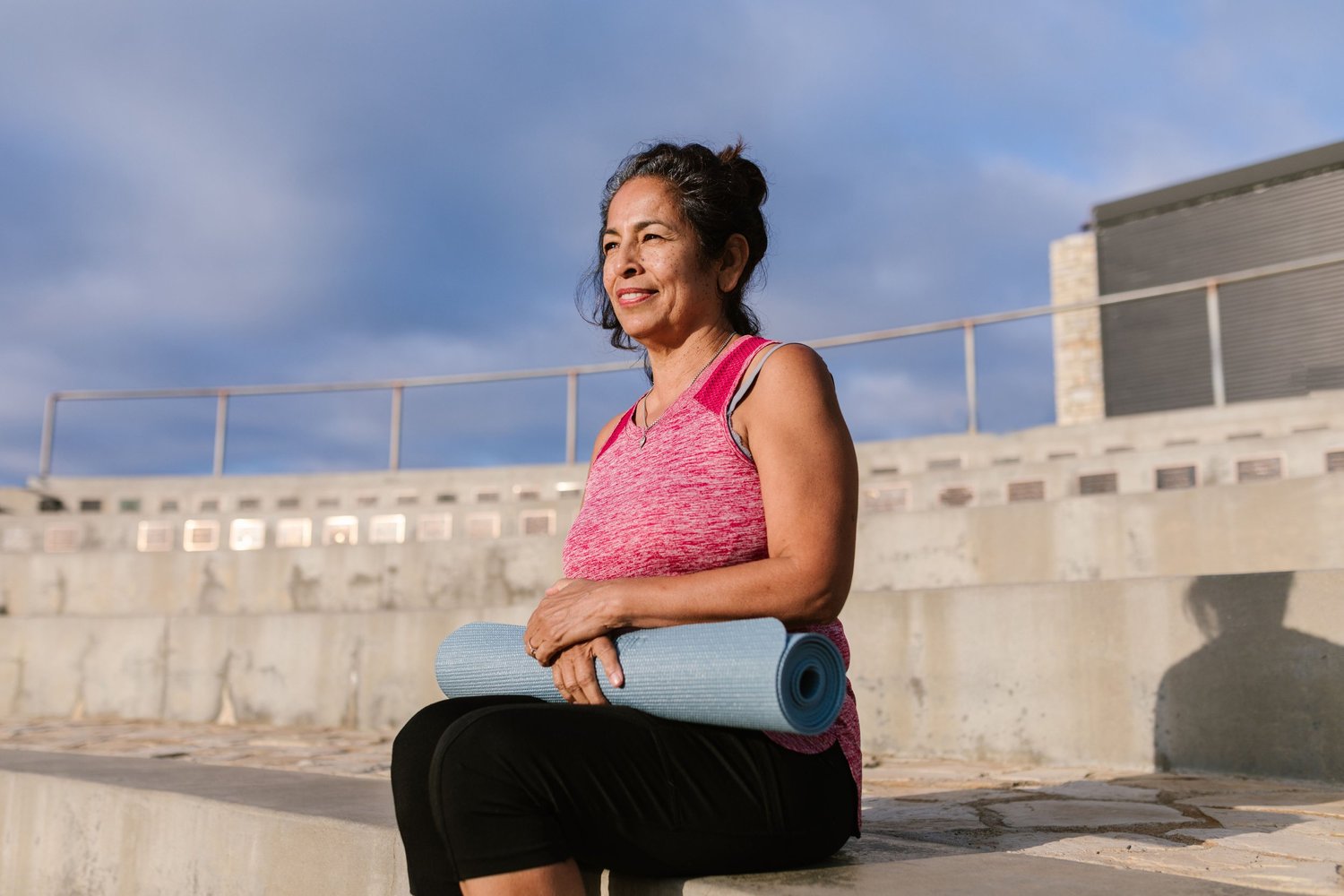A smiling woman sitting outdoors holding a rolled yoga mat