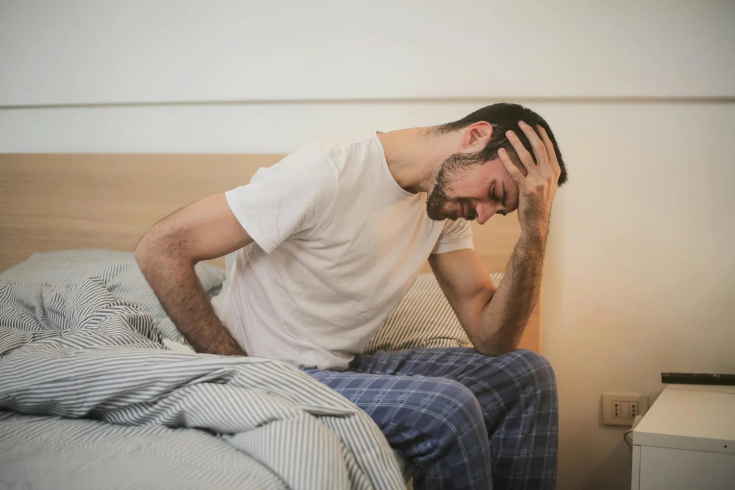 A man sitting on the edge of his bed, holding his head and side