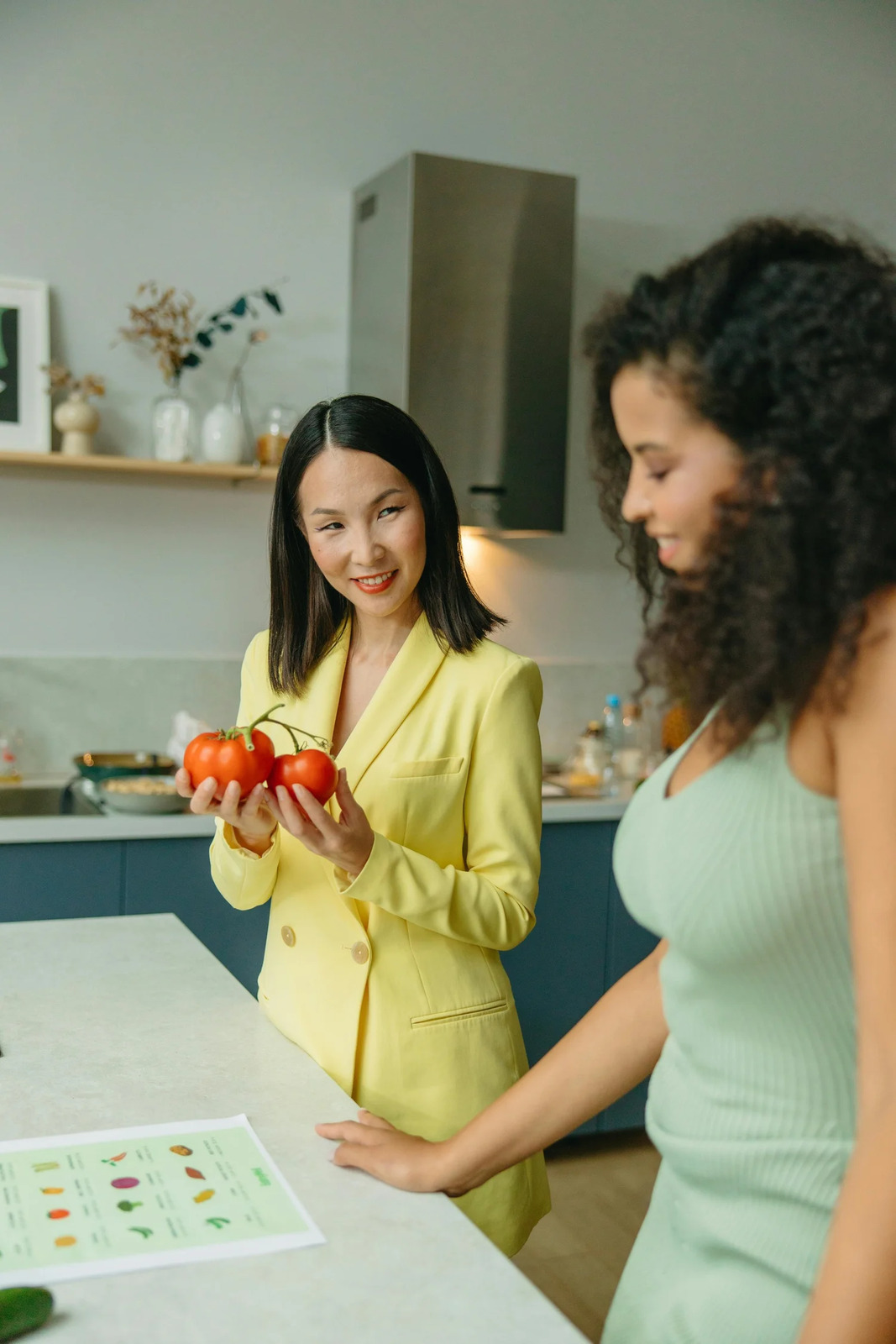 Two women talking in a kitchen, one holding tomatoes
