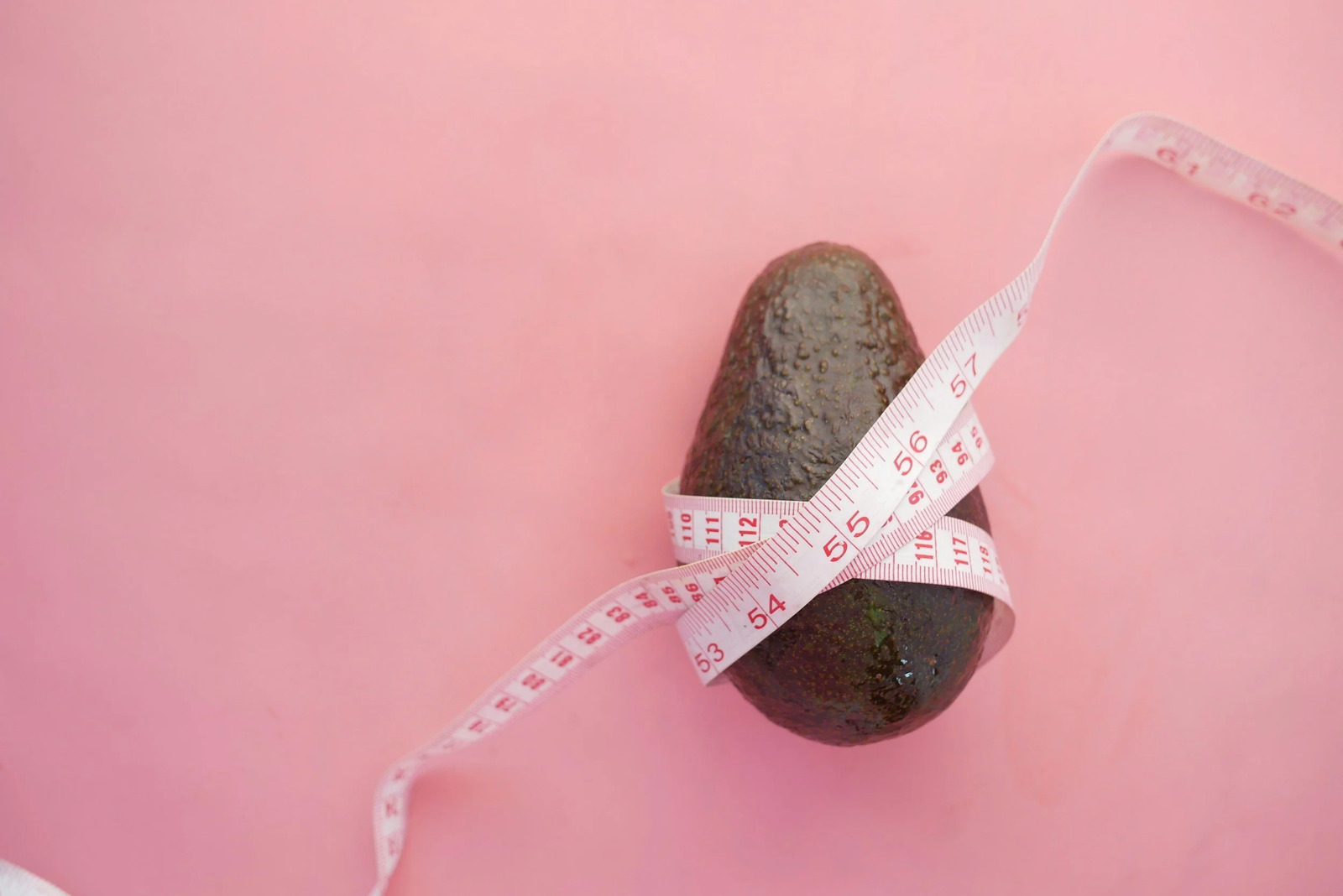 An avocado wrapped in a measuring tape on a pink background