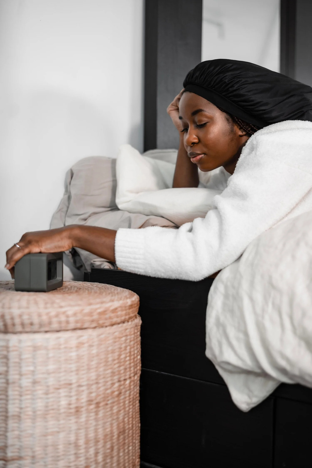 A woman sitting on the edge of her bed reaching for a bedside clock