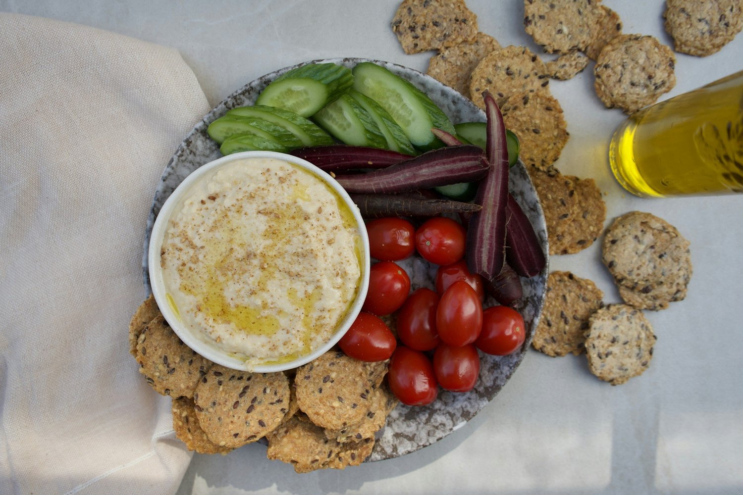 A plate of hummus with cucumber, tomatoes, purple carrots and seeded crackers