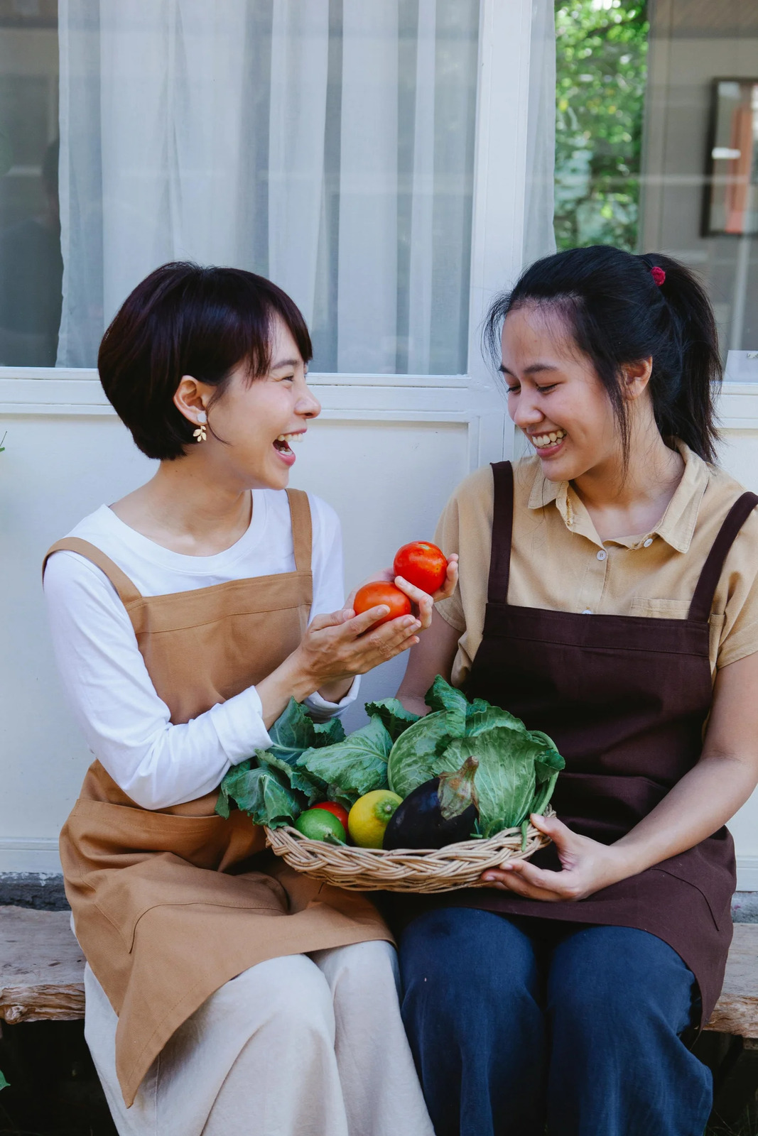 Two women in aprons laughing over a basket of fresh vegetables