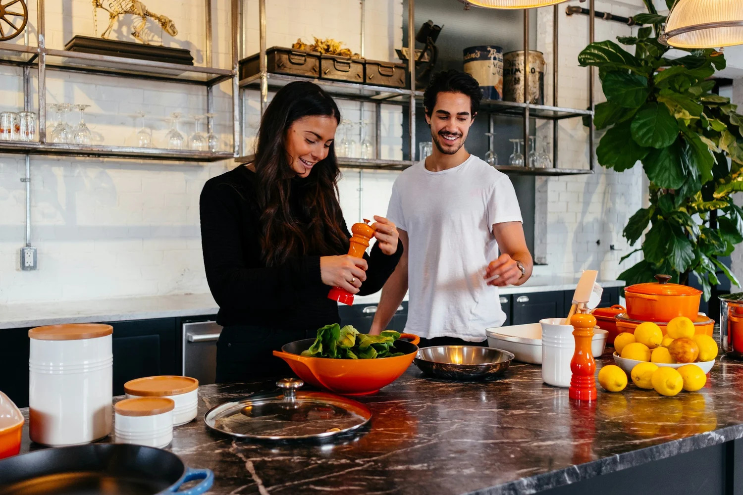 A young couple cooking together at a marble kitchen island, seasoning a bowl of greens