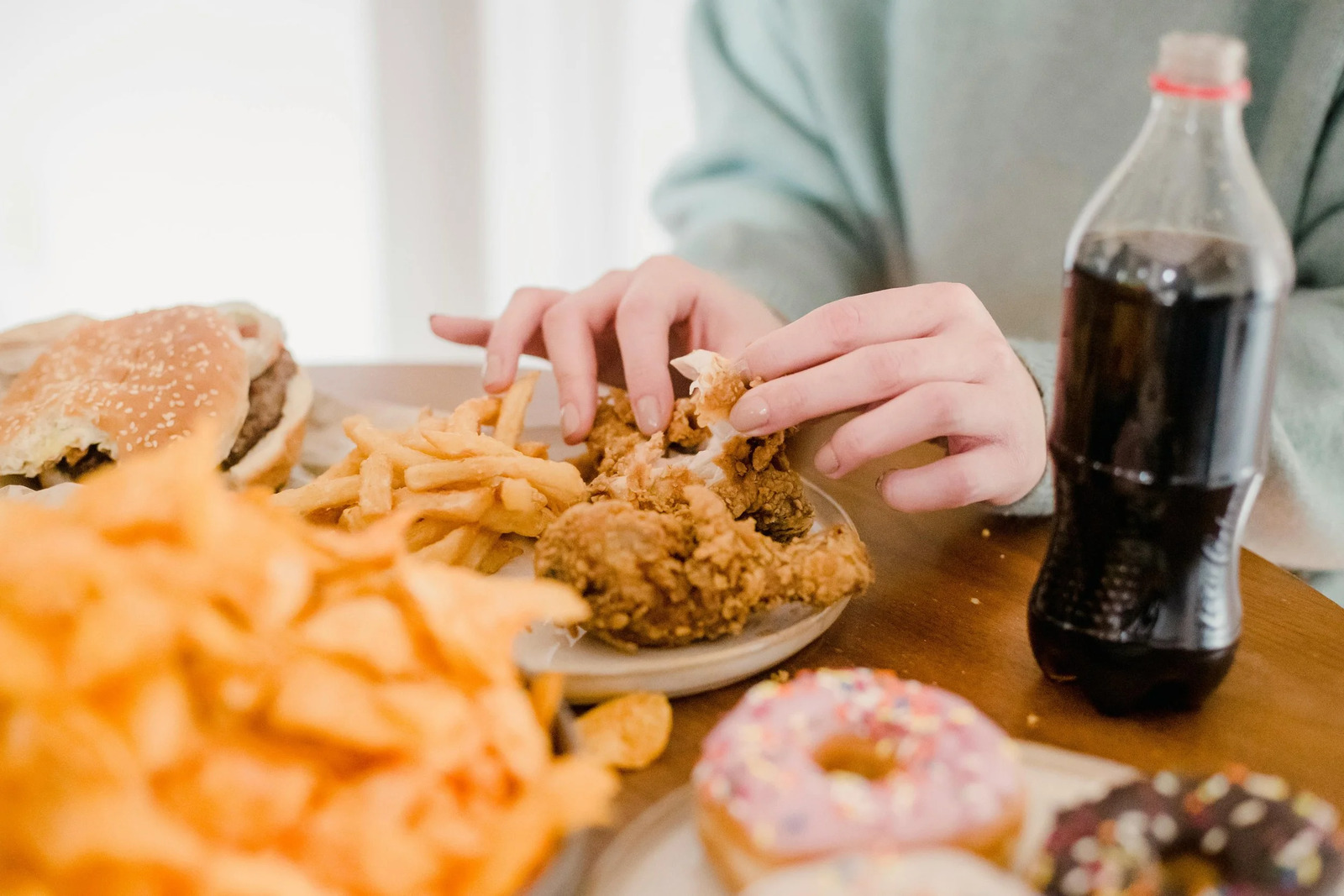 Hands reaching for fried chicken beside fries, doughnuts and soda