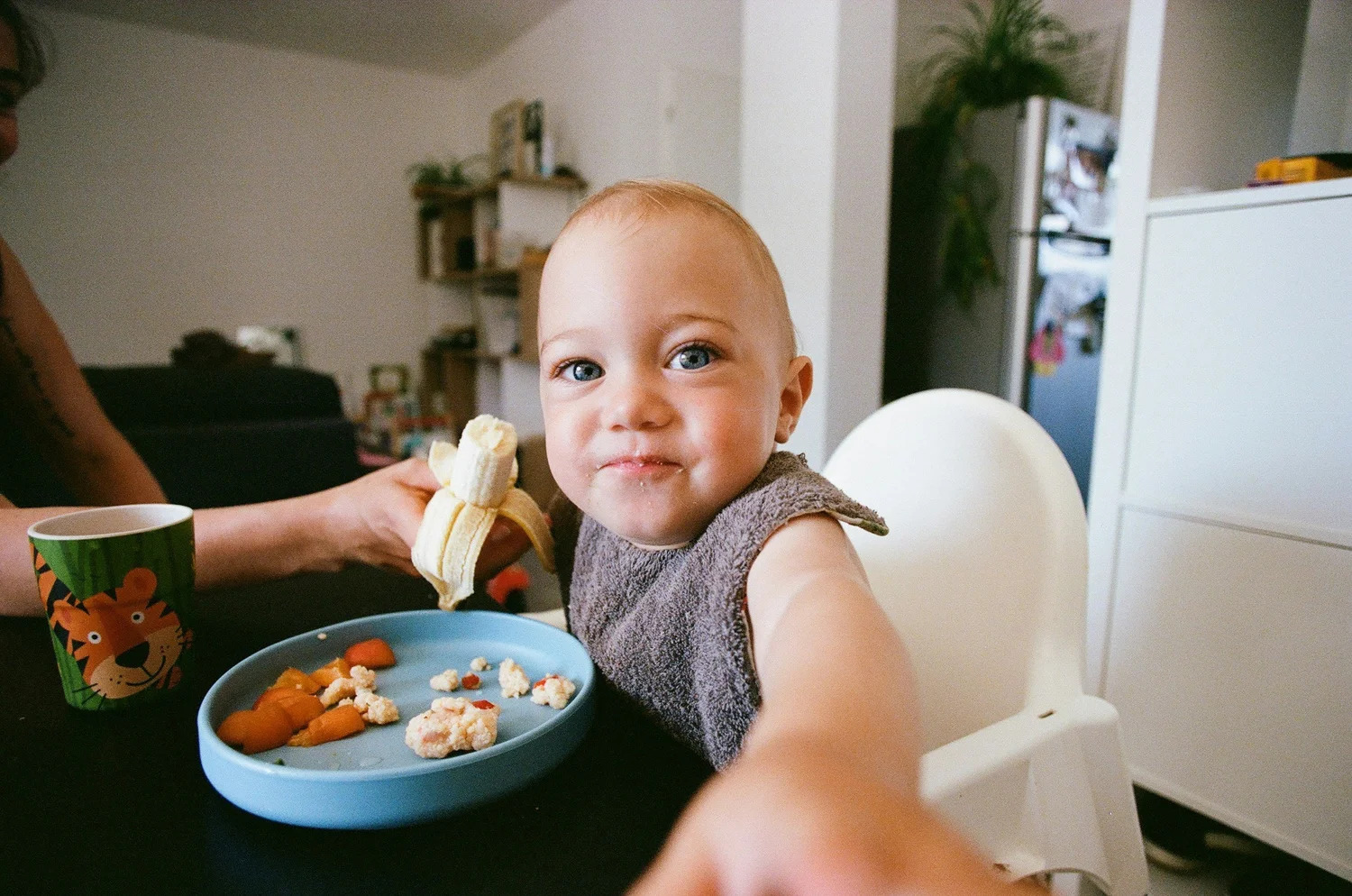 A smiling toddler in a high chair holding out a piece of food