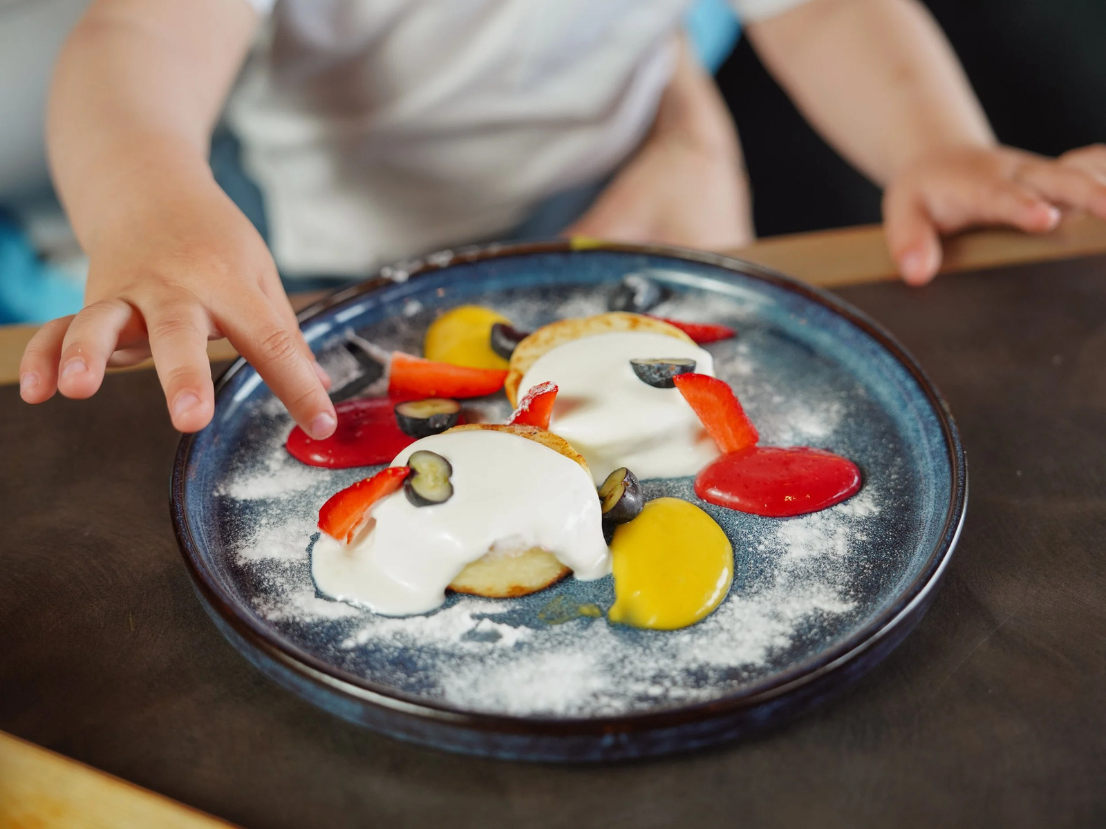 A child's hand reaching toward a plate of fruit and yogurt