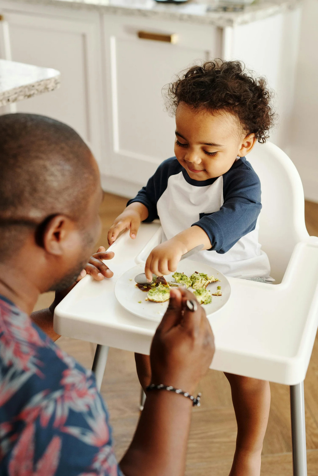 A toddler reaching for food on his high chair tray while his father watches