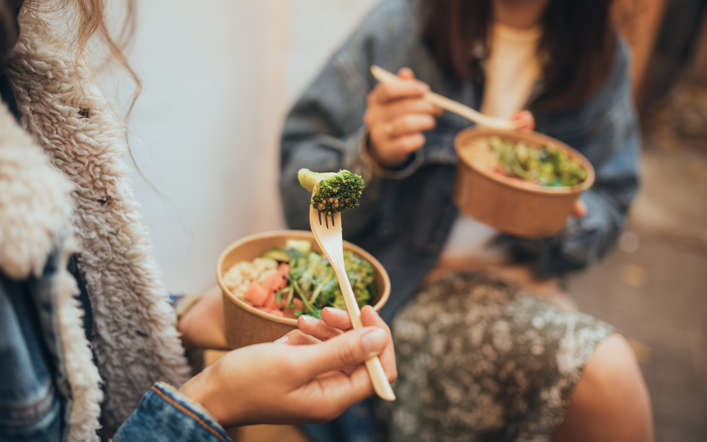 Two people sitting side by side outdoors eating grain bowls from takeaway containers, one lifting a floret of broccoli on a wooden fork, their faces out of frame