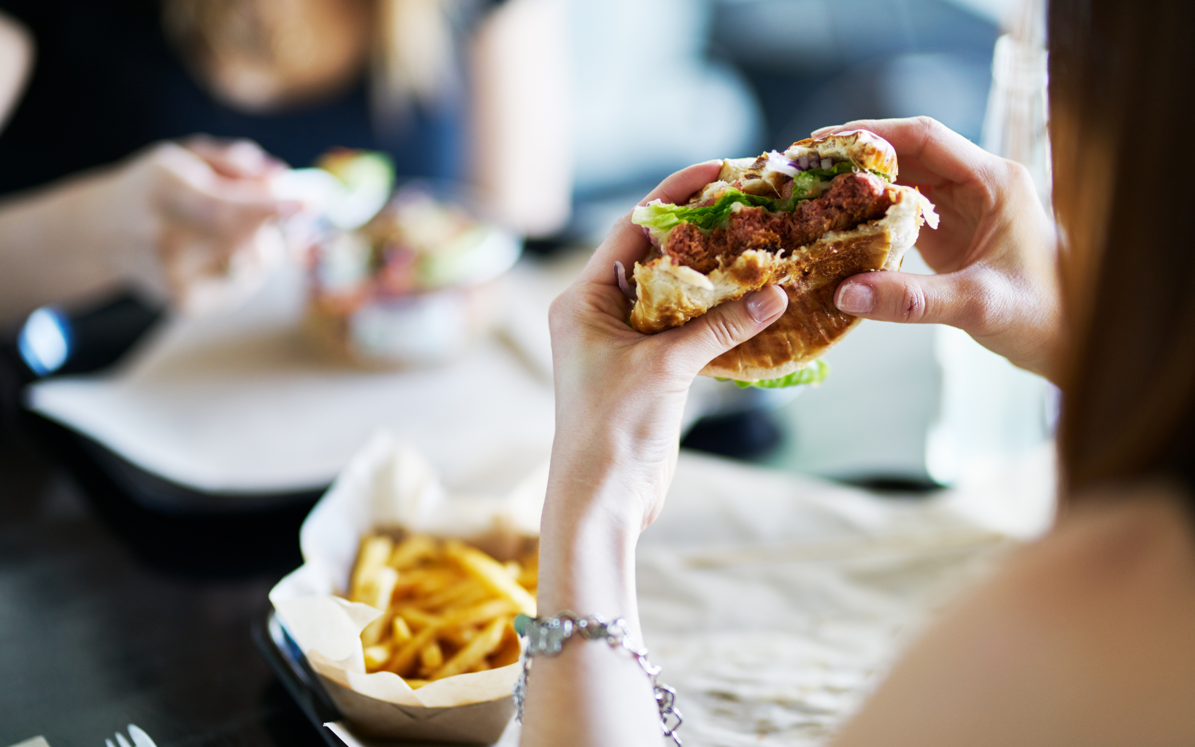 A person holding a partially eaten veggie burger with lettuce and red onion at a dark wooden table, with a kraft paper boat of golden fries beside them and a second person's hands reaching toward a salad bowl across the table