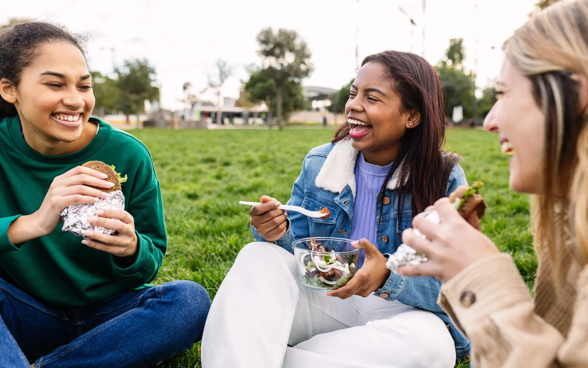 Three young women laughing together on a park lawn, eating wrapped sandwiches and a bowl of salad