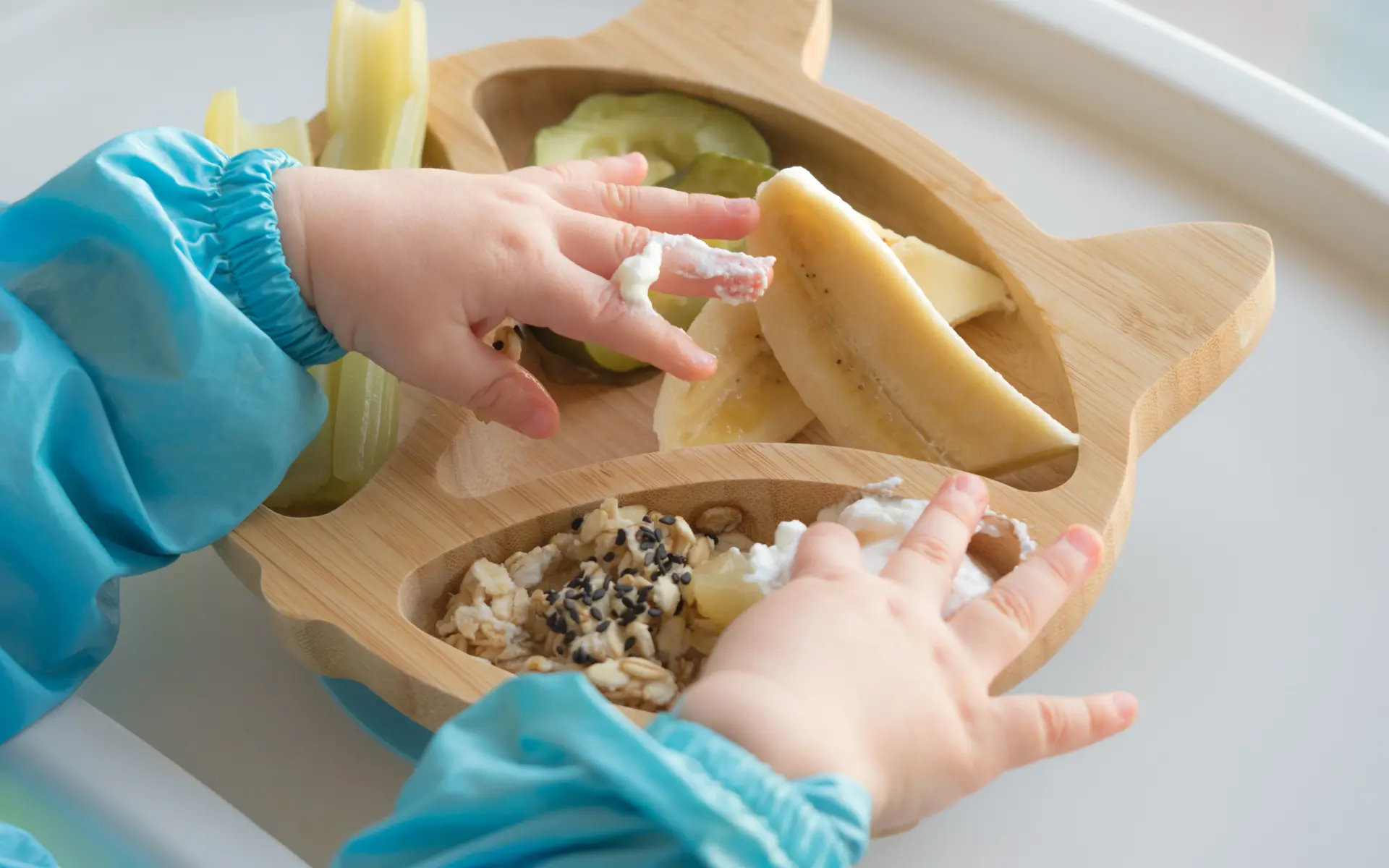 A baby’s hands reaching into a shaped bamboo feeding plate holding banana slices, cucumber rounds, celery sticks, and oats with yogurt