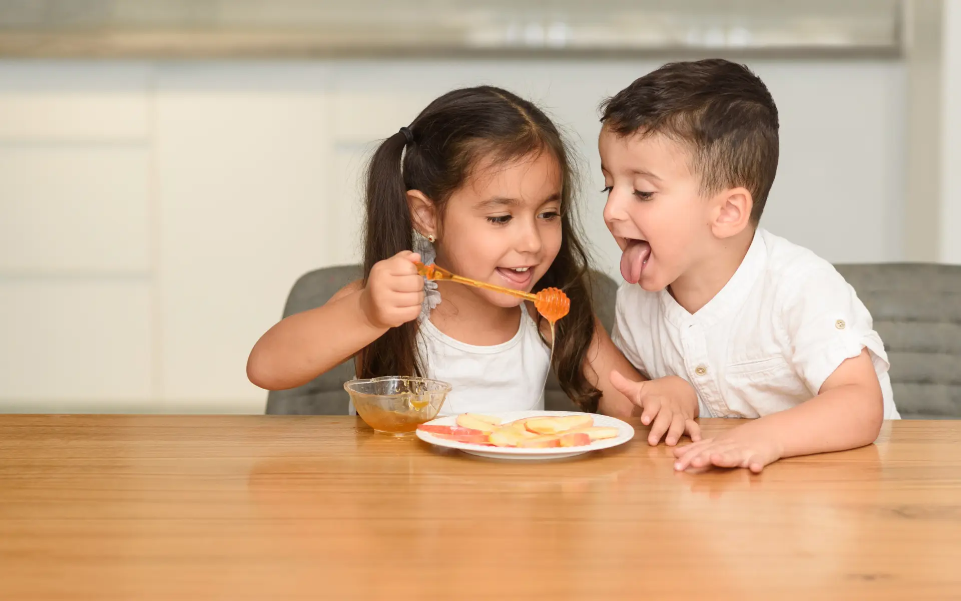 A young girl offering a honey dipper to a young boy at a wooden table with a plate of fruit