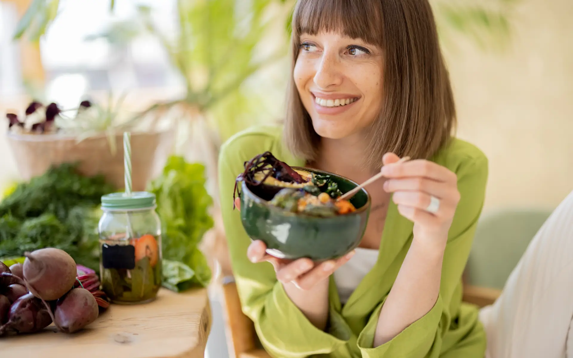 A young woman with a brown bob smiling as she holds a dark green bowl of salad at a light wooden table, with raw beets, fresh herbs, and a jar of infused water beside her