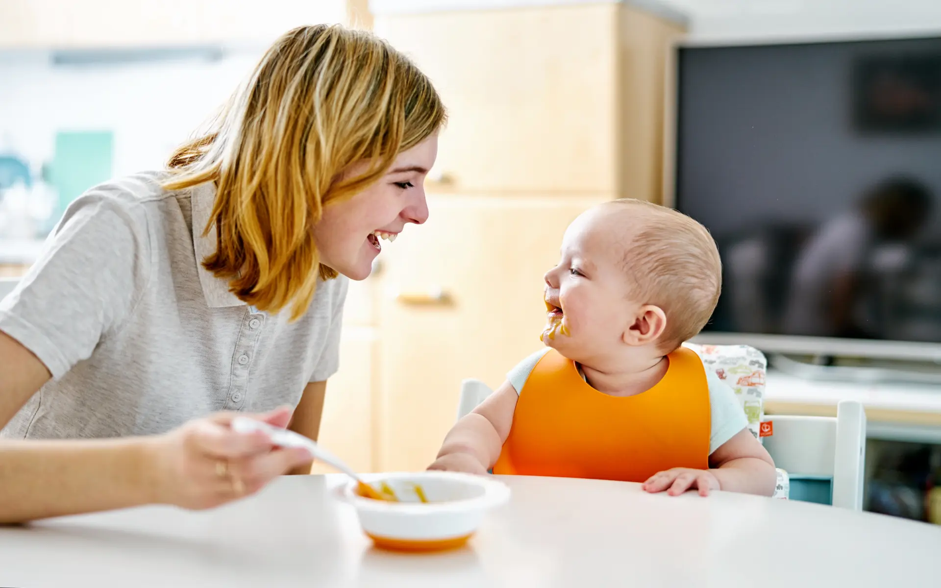 A mother laughing as she feeds spoonfuls of orange puree to her baby, who sits in a white high chair wearing a bright orange bib at a white table