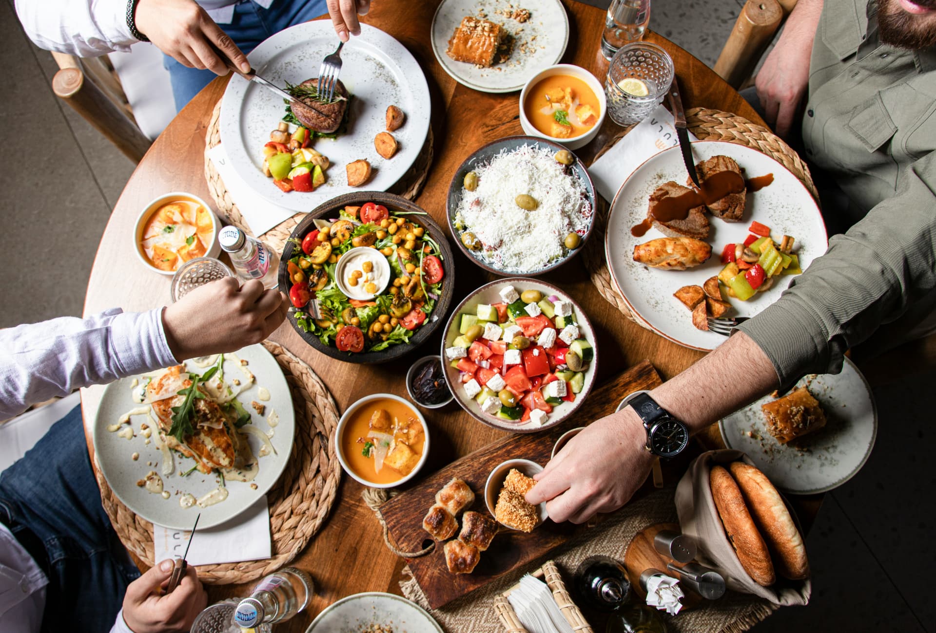 An overhead view of friends sharing a meal at a round wooden table, with hands reaching for Greek salad, a chickpea salad, soups, grilled meats, chicken, cheese pastries, and fresh bread