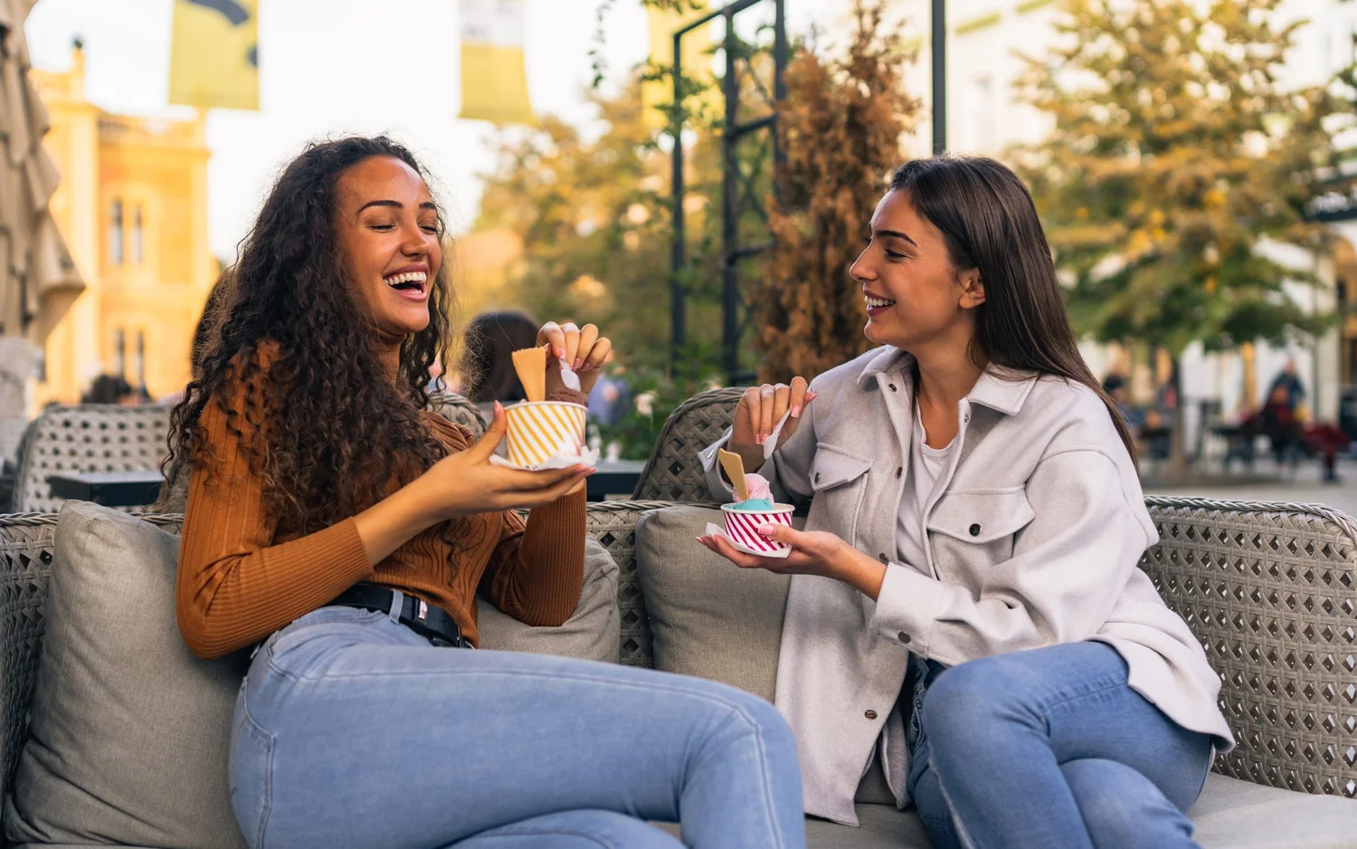Two women laughing over cups of frozen yogurt on an outdoor cafe sofa
